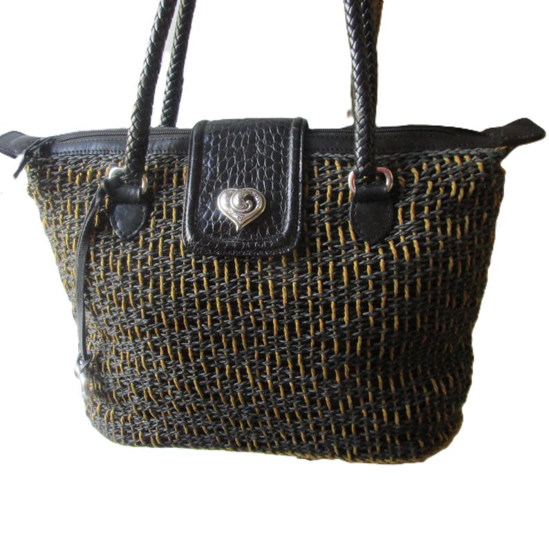 May include: Woven tote bag with black and gold threads. Features a black leather flap with a silver heart-shaped clasp. The bag has braided black handles and a zippered top closure.