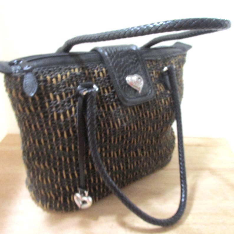 May include: A woven handbag with a black braided handle and accents. The bag features a black leather flap with a silver heart-shaped emblem. The body of the bag is a mix of black and tan woven material.