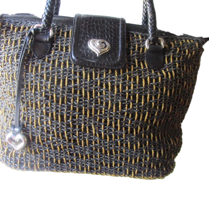May include: Woven handbag with a black and gold pattern. The bag features a black leather flap with a silver heart-shaped clasp. It has braided handles and a silver heart charm attached to a leather strap.