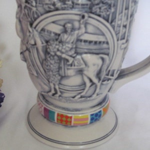 May include: A white ceramic stein with a detailed relief design of a horse and rider. The stein has a handle and a colorful base.