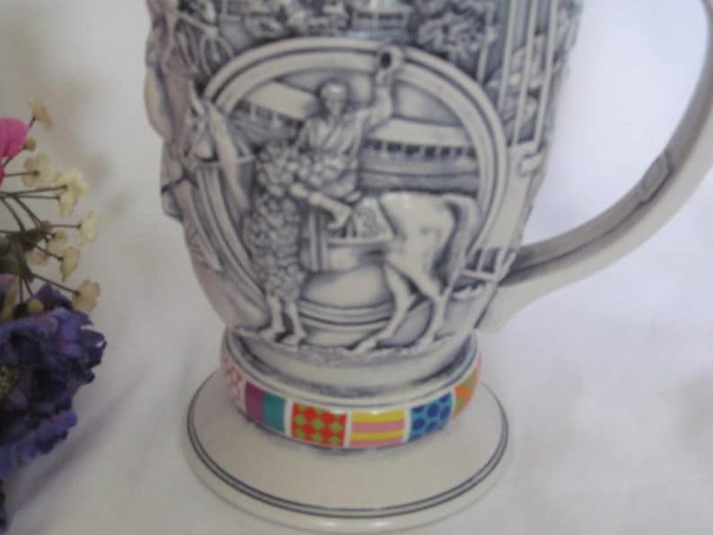 May include: A white ceramic stein with a detailed relief design of a horse and rider. The stein has a handle and a colorful base.