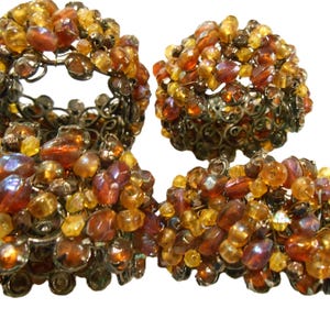 May include: A set of four circular napkin rings featuring a dark metal wire frame densely covered in a cluster of amber, orange, and golden-brown glass beads. The beads vary in size and shape, creating a textured, multi-faceted surface.
