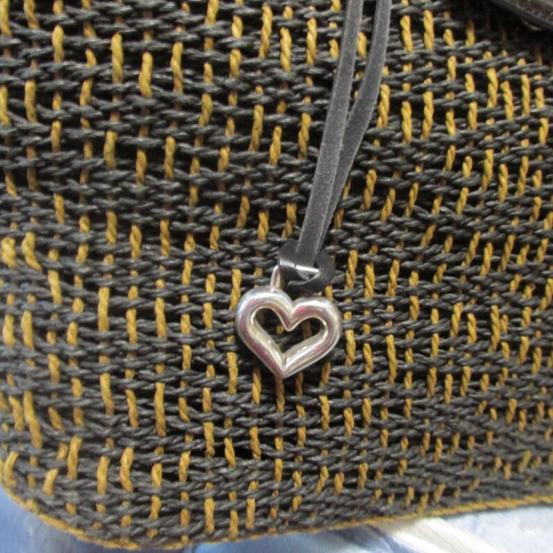 May include: Close-up of a woven handbag with a black and gold pattern. A silver heart charm hangs from a black strap. The bag's texture is visible, showcasing the intricate weave.