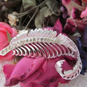 May include: A silver feather-shaped brooch with clear rhinestones. The brooch is set against a background of pink and purple fabric flowers.