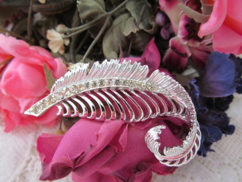 May include: A silver feather-shaped brooch with clear rhinestones. The brooch is set against a background of pink and purple fabric flowers.