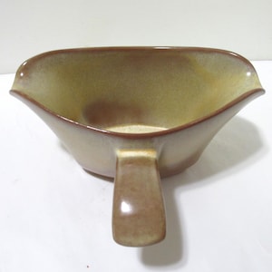 May include: A ceramic gravy boat featuring a smooth, rounded shape with a wide pouring spout and a single handle. The exterior and interior display a gradient color scheme, transitioning from a deep brown rim to a lighter, muted tan center.