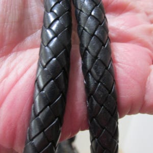 May include: Close-up of two black braided leather cords. The cords are thick and appear to be made of high-quality leather. The braiding is tight and even, creating a textured surface. The cords are likely part of a larger item, such as a belt or a bag strap.