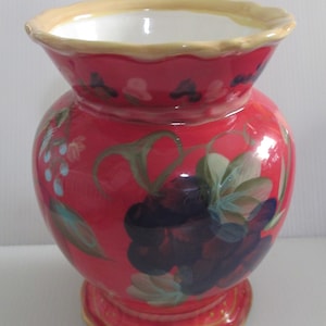 May include: A red ceramic vase with a yellow rim and a floral design featuring grapes and leaves. The vase has a scalloped rim and a rounded base.