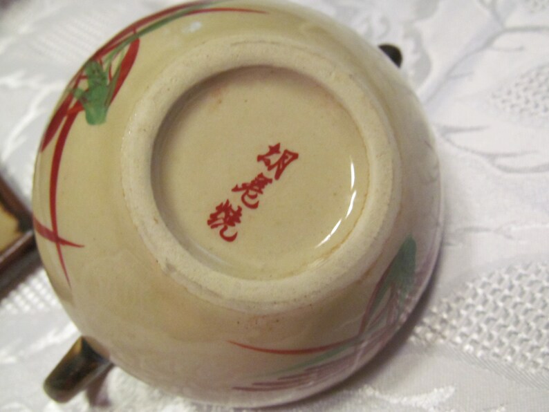 May include: A beige ceramic teapot with red and green floral designs. The bottom of the teapot has a red stamp that reads "華麗" in Japanese.