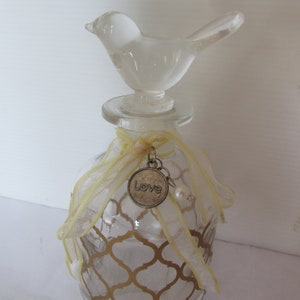 May include: A clear glass bottle with a gold geometric pattern and a clear glass bird on top. The bottle has a yellow ribbon with a charm that says "Love" hanging from it.