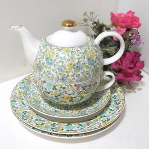 May include: A white porcelain tea set with a floral pattern in blue, yellow, and pink. The set includes a teapot, teacup, and two saucers. The teapot has a gold-colored knob on the lid. The set is arranged on a white surface.