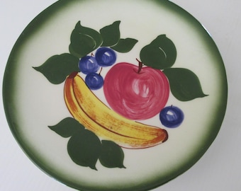 May include: A white ceramic plate with a green rim features a hand-painted design of a banana, apple, and blueberries with green leaves.