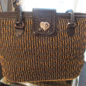 May include: Woven tote bag in a brown and tan color scheme. The bag features a textured weave pattern, a dark brown leather flap with a silver heart-shaped clasp, and braided handles. The bag has a zipper closure.