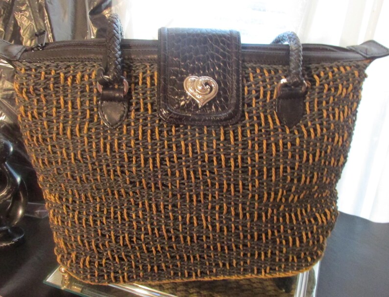 May include: Woven tote bag in a brown and tan color scheme. The bag features a textured weave pattern, a dark brown leather flap with a silver heart-shaped clasp, and braided handles. The bag has a zipper closure.