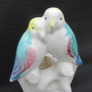 May include: A white ceramic figurine of two birds perched together. The birds have blue, yellow, and red accents on their wings and heads. The figurine is a decorative item, possibly a vase or ornament.