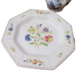 May include: White octagonal plate with a floral design in the center and around the edges. The flowers are in shades of blue, yellow, red, and purple. The plate has a ribbed texture and a scalloped edge. A blue and white cup is in the background.