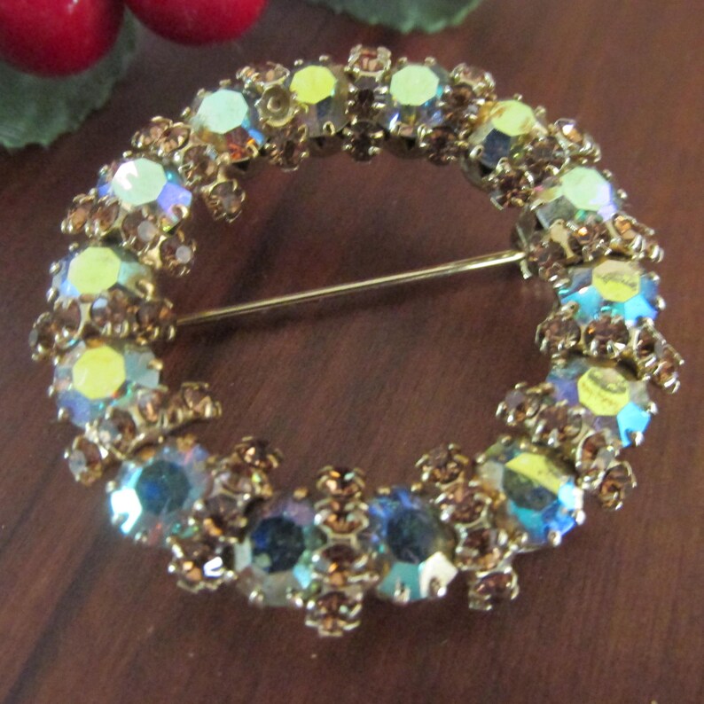 K&ouml;nnte beinhalten: Vintage, kreisf&ouml;rmige Brosche mit goldfarbenem Metallrahmen. Die Brosche ist mit schillernden, mehrfarbigen Strasssteinen in Blau-, Gelb- und Braunt&ouml;nen verziert. Ein klassisches Schmuckst&uuml;ck.