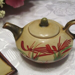 May include: A small, cream-colored teapot with a lid and a brown handle. The teapot is decorated with red and green floral designs.