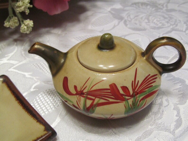 May include: A small, cream-colored teapot with a lid and a brown handle. The teapot is decorated with red and green floral designs.