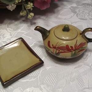May include: A small, beige teapot with a lid and a brown handle. The teapot has a floral design with red and green accents. A small, square, beige plate with a brown rim is also pictured.