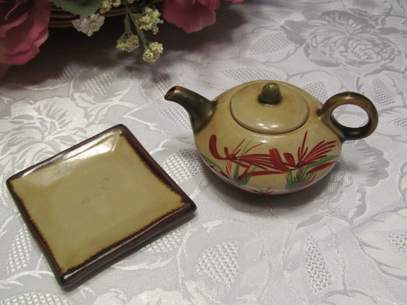 May include: A small, beige teapot with a lid and a brown handle. The teapot has a floral design with red and green accents. A small, square, beige plate with a brown rim is also pictured.