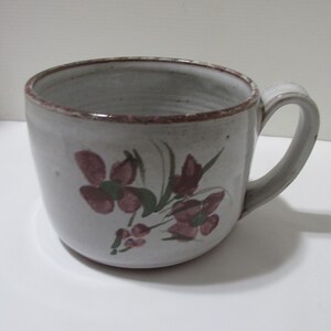 May include: A ceramic mug with a speckled light gray finish and a hand-painted floral design in muted red and green. The mug features a wide, rounded body, a sturdy side handle, and a dark red rim detail that complements the painted flower motif.