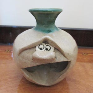 May include: A beige ceramic vase with a speckled texture and a green glazed neck. The body of the vase features a sculpted face with two round eyes, a prominent nose, and a wide, open mouth cavity that serves as an opening in the front of the vessel.