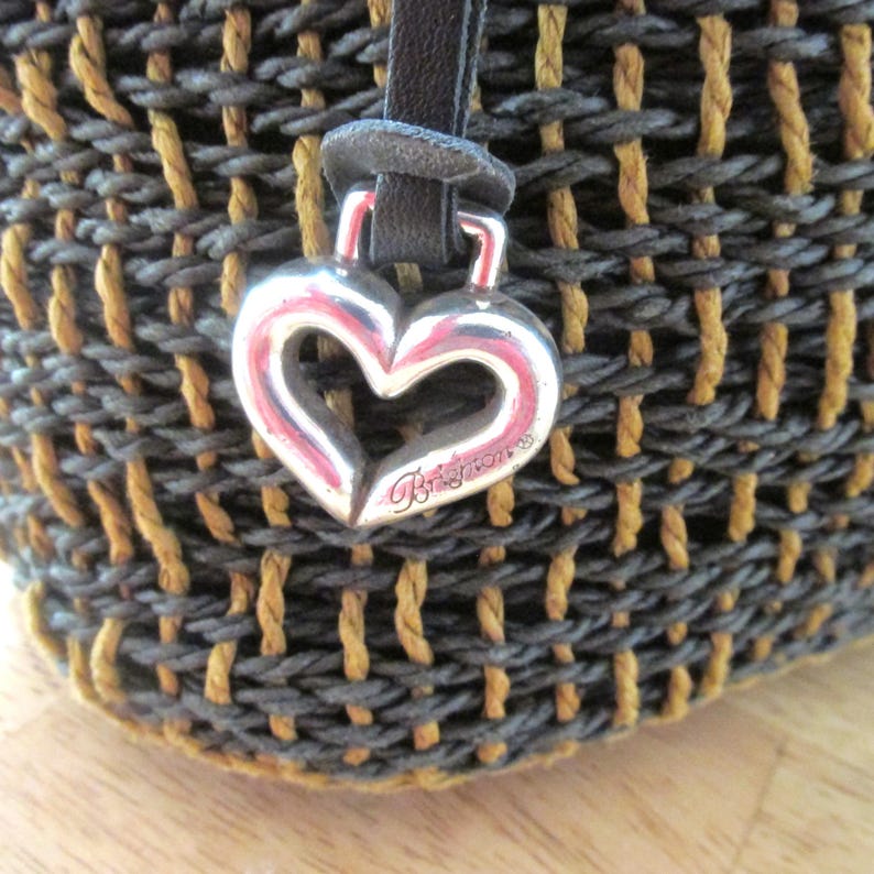May include: Close-up of a woven handbag with a silver heart-shaped charm. The bag features a mix of gray and tan woven material. The heart charm is engraved with the brand name "Brighton" in cursive.