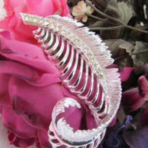 May include: A silver feather-shaped brooch with rhinestones set against a background of pink and purple fabric flowers.