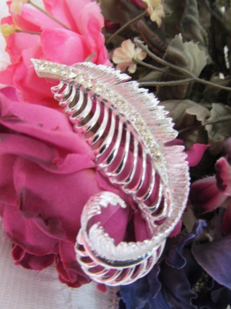 May include: A silver feather-shaped brooch with rhinestones set against a background of pink and purple fabric flowers.
