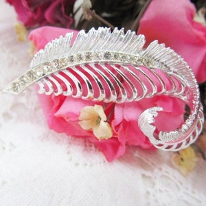 May include: A silver feather-shaped brooch with clear rhinestones. The brooch is set against a background of pink fabric.