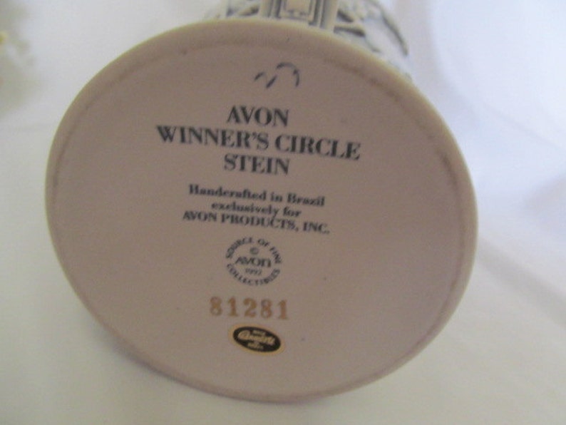 May include: White ceramic stein with a black and white image of a building. The bottom of the stein has the text "AVON WINNER'S CIRCLE STEIN" and "Handcrafted in Brazil exclusively for AVON PRODUCTS, INC." along with a logo and the number 81281.