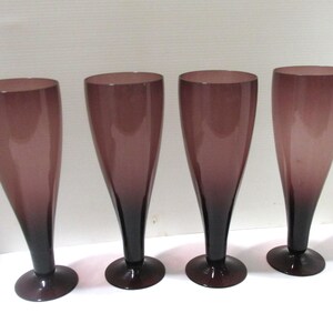 May include: A set of four tall, slender drinking glasses with a smooth, tapered silhouette. Each glass features a deep purple gradient that transitions from a lighter shade at the rim to a darker, more saturated purple hue at the base and stem.