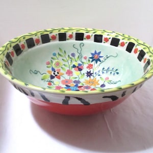 May include: A decorative ceramic bowl with a floral design. The bowl features a light green interior with colorful flowers and a red exterior. The rim has a black and white pattern with a yellow border.