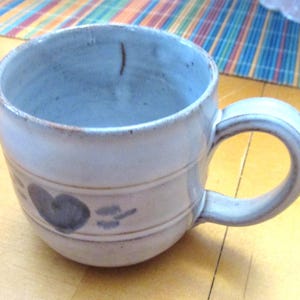May include: A light gray ceramic mug with a rounded body and a sturdy handle. The exterior features a horizontal band embossed with a simple dark gray heart shape and small decorative marks. The glaze has a subtle, uneven texture throughout.
