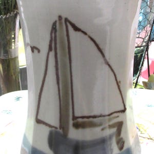 May include: A white ceramic vase with a hand-painted sailboat design. The vase features a brown outline of a sailboat with white sails and a blue water line. The vase is approximately 8 inches tall.