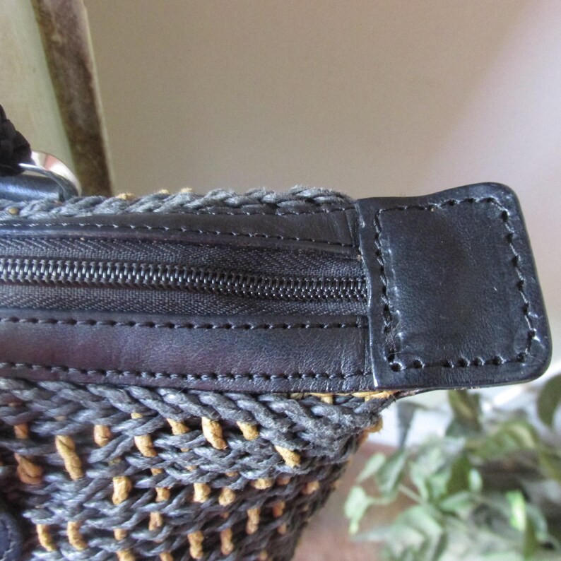 May include: Close-up of a woven handbag with a black zipper and leather trim. The bag features a gray and tan woven pattern, with a black leather accent on the zipper area. The stitching is visible, adding detail to the design.