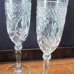 May include: Two clear crystal champagne flutes with intricate diamond and leaf-like cut patterns. The flutes have a round base, a slender stem, and a detailed design on the bowl. The glasses are displayed on a wooden surface.