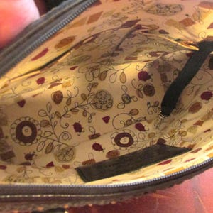 May include: Interior view of a dark brown handbag. The lining is a cream-colored fabric with a floral and geometric pattern in shades of brown, red, and yellow. A black strap and zipper are visible.