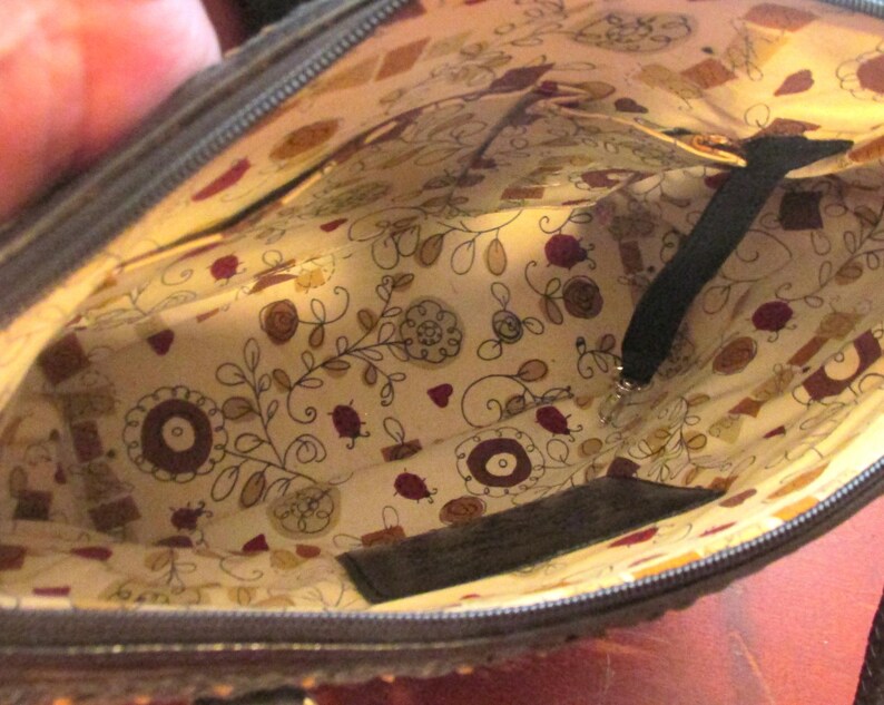 May include: Interior view of a dark brown handbag. The lining is a cream-colored fabric with a floral and geometric pattern in shades of brown, red, and yellow. A black strap and zipper are visible.