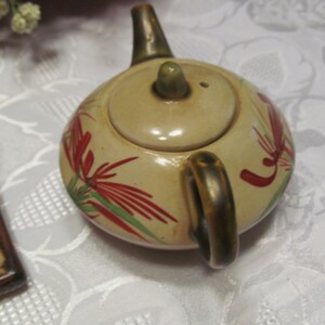 May include: A small, cream-colored teapot with a gold handle and lid. The teapot is decorated with red and green floral designs.