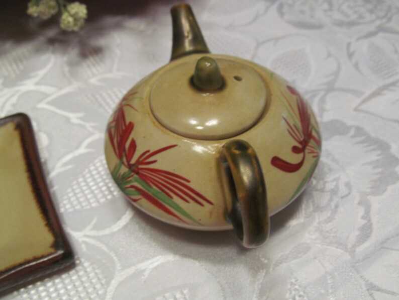 May include: A small, cream-colored teapot with a gold handle and lid. The teapot is decorated with red and green floral designs.