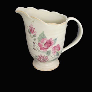 May include: A cream-colored ceramic pitcher with a floral design. The pitcher has a scalloped rim and a handle. The flowers are pink and green.