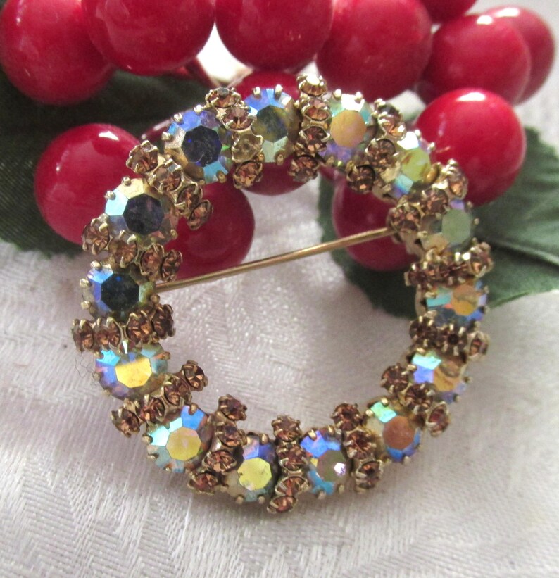 K&ouml;nnte beinhalten: Vintage, kreisf&ouml;rmige Brosche mit irisierenden Kristallen in Blau, Gelb und Gr&uuml;n, gefasst in einem goldfarbenen Metallrahmen. Die Brosche hat einen Nadelverschluss und ist von kleinen, braunen Kristallen umgeben. Ein klassisches Schmuckst&uuml;ck.