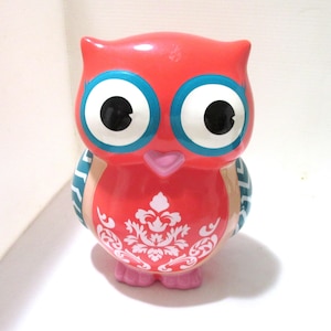 May include: A ceramic owl figurine featuring a bright red body with a white damask pattern on the chest. The owl has large white eyes with blue rims, black pupils, a small pink beak, and pink feet. The wings are decorated with a blue and white chevron pattern.