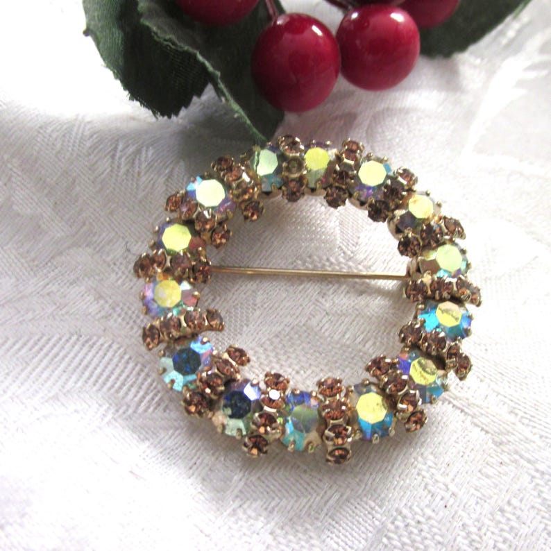K&ouml;nnte beinhalten: Vintage, kreisf&ouml;rmige Brosche mit goldfarbenem Metallsockel. Die Brosche ist mit schillernden, mehrfarbigen Strasssteinen und kleineren braunen Strasssteinen verziert. Ein klassisches Schmuckst&uuml;ck.