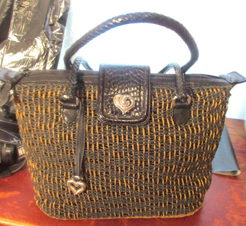 May include: A woven handbag with a black and tan pattern. The bag has a braided handle and a black flap with a silver heart-shaped emblem. A black leather tag with a silver heart charm hangs from the zipper.