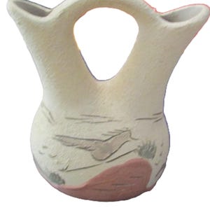 May include: A cream-colored, handcrafted pottery vase with a distinctive handle and two spouts. The vase features hand-painted designs in shades of brown, pink, and green. The vase has a rounded body and a unique shape, perfect for home decor.