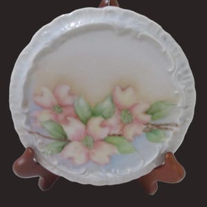 May include: A round, white ceramic plate with a scalloped edge, featuring a hand-painted floral design. The flowers are pink with green centers and leaves, set against a soft, gradient background. The plate is displayed on a brown wooden stand.