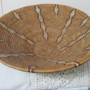 May include: A round, woven basket with a spiral center and radiating patterns. The basket is primarily light brown with accents of darker brown and cream-colored stripes. The rim is reinforced with a darker brown band.